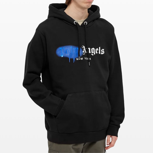 A person wears a Palm Angels New York Sprayed Logo Hoodie, showcasing the brand's signature blue and white sprayed logo on the front, which gives off a graffiti-style vibe. Featuring a drawstring and kangaroo pocket, the black hoodie adds a stylish touch as they stand against a plain background.