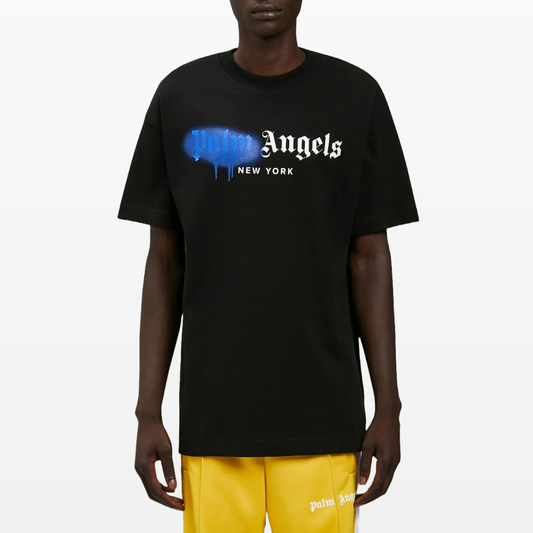 Dressed in a Palm Angels New York Sprayed Logo T-Shirt featuring the "Palm Angels New York" lettering in white and blue, an individual pairs it with yellow shorts adorned with the iconic "Palm Angels" text near the hem. The plain white background subtly enhances this streetwear ensemble's sleek, modern vibe.