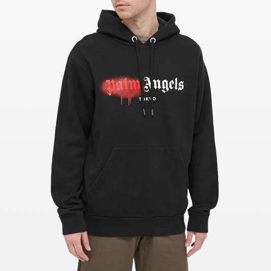 A person is dressed in a Palm Angels Tokyo Sprayed Logo Hoodie, crafted from premium heavyweight cotton, showcasing "Palm Angels" in striking red and white text across the chest. They have stylishly paired it with brown pants that include drawstrings and a front pocket for an extra touch of flair.