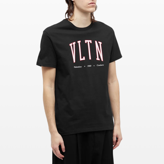 A person wearing a black Valentino VLTN College Logo T-Shirt, emblazoned with "VLTN" in pink letters and featuring "Valentino 1960 Couture" below. This luxury streetwear item is paired with black pants, complementing their shoulder-length hair for a chic and modern appearance.