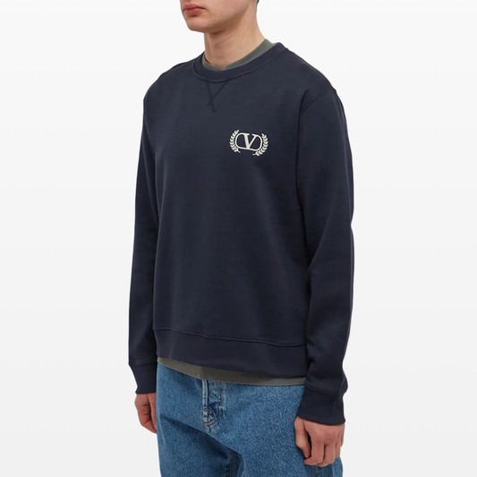 A person wearing a Valentino Varsity V Logo Sweatshirt in navy blue, showcasing understated embroidered branding on the upper left chest, stands against a plain white background. The outfit is complemented by light blue jeans, but the person's face is not visible in the image.