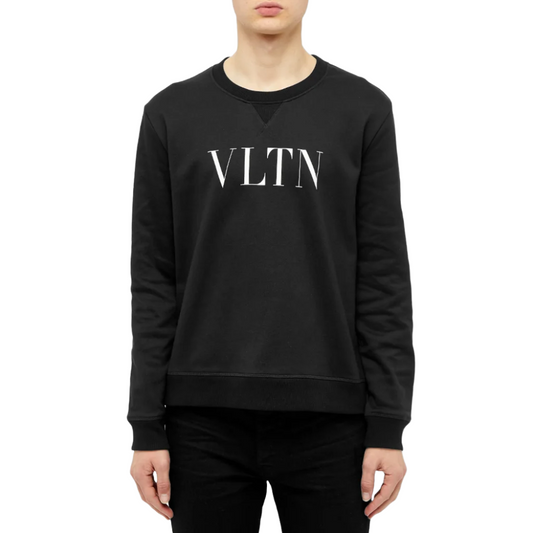 A person wears the Valentino VLTN Logo Sweatshirt, showcasing the "VLTN" branding in white across the front. Manufactured in Italy, this outfit is paired with black pants against a plain white backdrop, highlighting its stylish appeal.