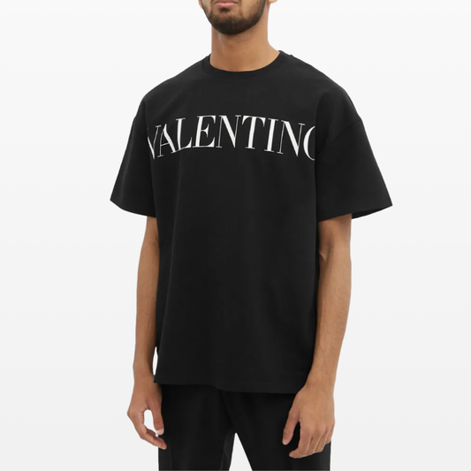 A person wearing a Valentino Printed Logo T-Shirt in black, crafted from premium cotton, featuring bold white letters stands against a plain white background.