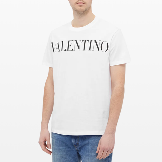 Someone is wearing a Valentino Printed Logo T-Shirt in white, made from 100% cotton, featuring "Valentino" prominently in bold black letters on the chest. When paired with blue jeans, this outfit creates a striking contrast against the plain white backdrop.