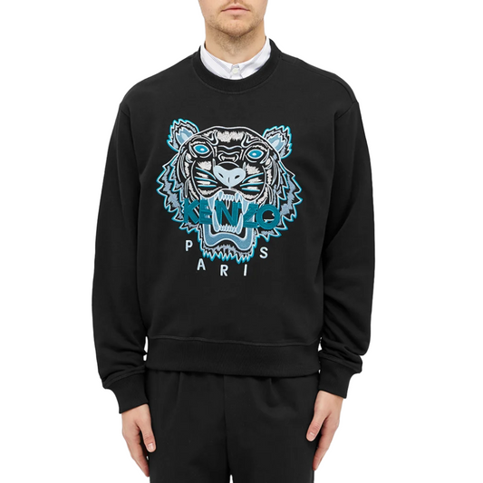 A man clad in a sleek black Kenzo Classic Tiger Embroidered Sweatshirt, showcasing the Parisian style with its iconic tiger emblem and bold "PARIS" lettering, stands against a plain white backdrop.