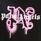 The Palm Angels Miami Monogram Sprayed Logo T-Shirt, featuring the "Palm Angels" logo in white gothic font against a black background, subtly displays "Miami Monogram" beneath. A striking pink graffiti-style "N" behind the text adds an edgy flair.
