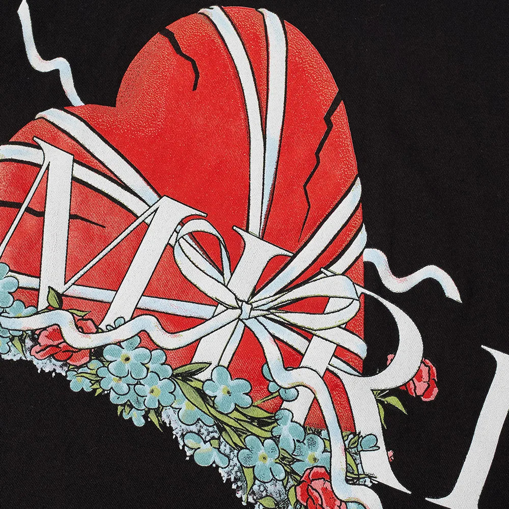 The Amiri Broken Heart T-Shirt by Amiri features a striking design of a cracked red heart entwined with white ribbons, accented with blue flowers. The letters "M" and "RI" appear in white against a black background, exuding elegance on 100% cotton fabric.