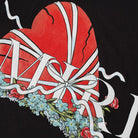 The Amiri Broken Heart T-Shirt by Amiri features a striking design of a cracked red heart entwined with white ribbons, accented with blue flowers. The letters "M" and "RI" appear in white against a black background, exuding elegance on 100% cotton fabric.