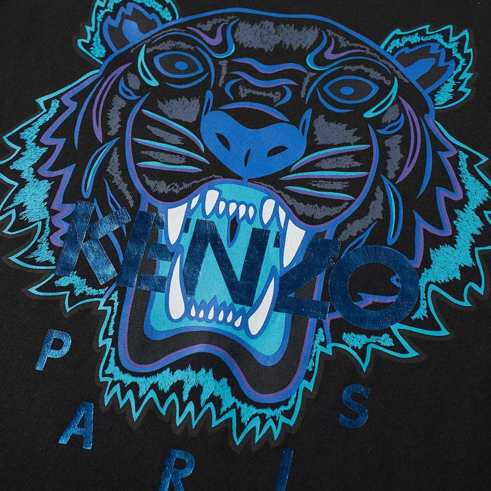 The Kenzo Holiday Capsule Tiger T-Shirt showcases an electric blue tiger face with sharp teeth in shades of blue and black, overlaid with the text "KENZO PARIS." The vibrant illustration stands out against the sleek black background of this streetwear piece from Kenzo.
